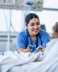 A female nurse of Middle Eastern decent sits at the edge of a hospital bed as she check in on her young patient. She is wearing blue scrubs and is attempting to cheer the young girl up.